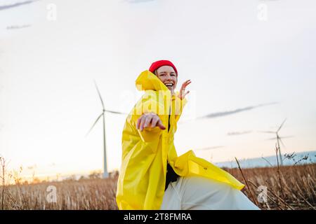 Una giovane donna gioiosa, con un impermeabile giallo e un berretto rosso vibrante, danzerà in un campo al tramonto con turbine eoliche sullo sfondo. Foto Stock