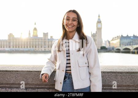 Una giovane donna allegra con un cappotto alla moda sorride mentre posa di fronte all'iconico Big Ben e alle Houses of Parliament di Londra durante una giornata di sole Foto Stock