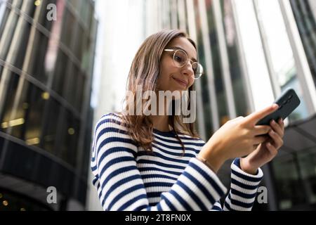 Una giovane donna sorridente in un top a righe usa il suo smartphone sullo sfondo di edifici moderni a Londra. Foto Stock