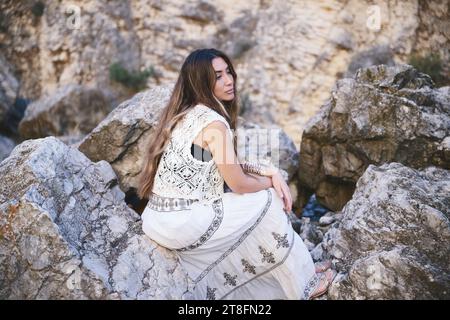 Vista laterale della donna in un abito bianco senza maniche adornato da motivi si siede su rocce aspre che guardano lontano con i capelli avvolti sul suo shou Foto Stock