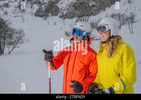 Una coppia allegra e piena di colorate attrezzature invernali si pone con gli sci su una montagna innevata, trasudando la gioia di una vacanza sulla neve. Foto Stock