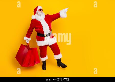 Foto a lunghezza intera del costume rosso di babbo natale vestito da nonno che dirige il negozio promozionale dello spazio delle dita isolato su sfondo giallo Foto Stock