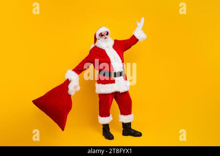 Ritratto a grandezza naturale dell'eccentrico sorridente babbo natale Hold di Capodanno presenta la borsa alzata a mano isolata su sfondo giallo Foto Stock