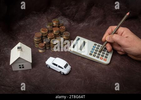 Mano che regge un pastello e calcola le tasse con la casa modello, l'auto e le pile multiple di monete su pelle marrone Foto Stock