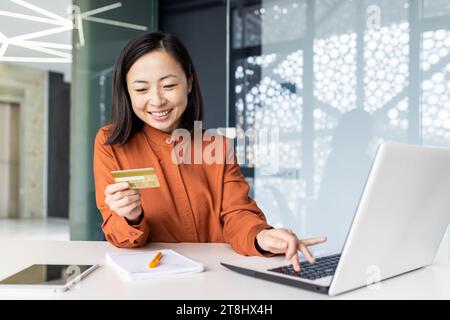 Ritratto di una giovane donna asiatica soddisfatta di successo all'interno dell'ufficio sul posto di lavoro, donna d'affari che sorride, che usa la carta di credito bancaria di debito per lo shopping online, che lavora con un computer portatile. Foto Stock