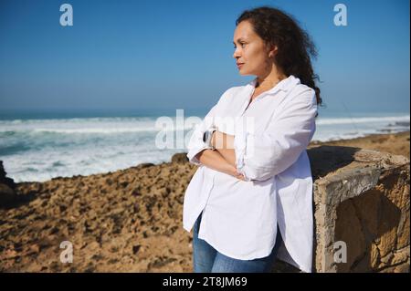 Bella donna pensiva di mezza età vestita con camicia casual bianca e jeans, in piedi con le braccia piegate sullo sfondo dell'Oceano Atlantico, sognante Foto Stock
