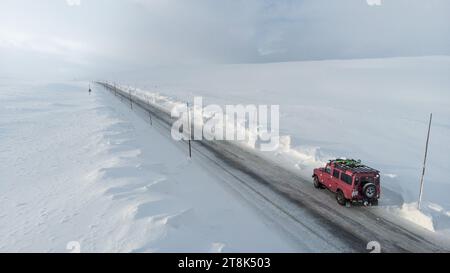 Una jeep rossa brillante che guida lungo un'autostrada innevata, con un grande camion sullo sfondo carico di neve Foto Stock