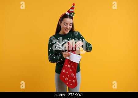 Felice giovane donna con maglione natalizio che prende il regalo da calza su sfondo arancione Foto Stock