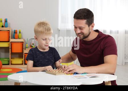 Sviluppo di competenze motorie. Padre felice che aiuta suo figlio a giocare con geoboard e fasce di gomma al tavolo bianco in camera Foto Stock