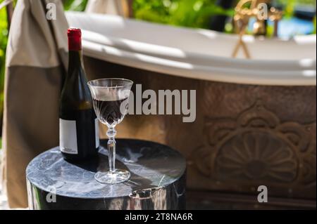 Un bicchiere di vino rosso con una bottiglia posta su un tavolo di marmo accanto alla vasca esterna per godersi un momento di relax. Foto Stock