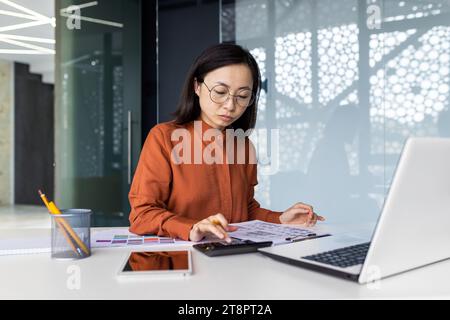 Sicura di sé, seria, concentrata e concentrata, donna che lavora in ufficio con la carta, donna con la calcolatrice che pensa di controllare i conti, relazioni finanziarie, asiatica con notebook che prepara documenti. Foto Stock