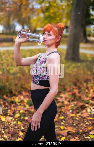 Redhead 20s donna caucasica che beve da una bottiglia dopo aver corso di allenamento in parco a fine autunno Foto Stock