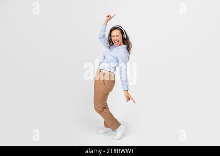 Allegra donna millenaria europea con cuffie wireless a bocca aperta, balla, ascolta musica e canta Foto Stock