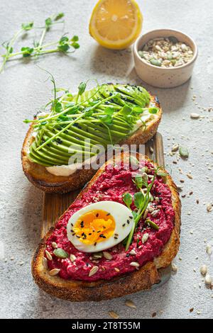 Pane tostato a grani interi con formaggio cremoso, avocado, hummus di barbabietole, semi, uova e microgreens (germogli di piselli germogliati) su fondo grigio chiaro. Sano f Foto Stock