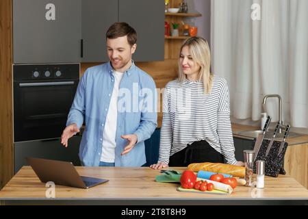 Piacevole coppia di famiglia in piedi vicino a un grande tavolo di legno in una cucina moderna, guardando lo schermo del computer portatile. Felice giovane coppia che inizia a preparare il pranzo Foto Stock