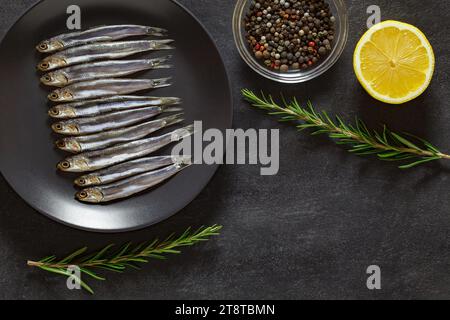 Acciuga europea, hamsa fresca, diversi piccoli pesci disposti in fila su piatto grigio, limone, rosmarino, mix di pepe, su fondo scuro, vista dall'alto, spazio per c Foto Stock