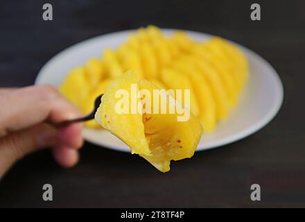 La forchetta di primo piano raccoglie un pezzo di ananas fresco con fette sfocate sullo sfondo Foto Stock