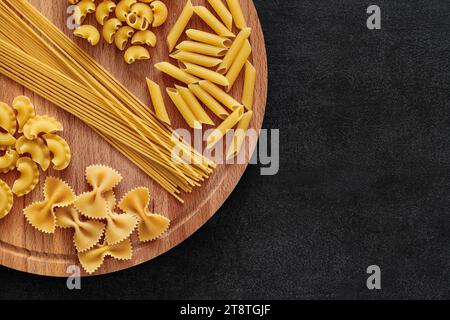 Pasta, mix di farfalle crude, spaghetti, rigatoni al pipa, maccheroni, penne rigate, cresta di gallo, su piedistallo rotondo in legno su sfondo scuro, vista dall'alto, s Foto Stock