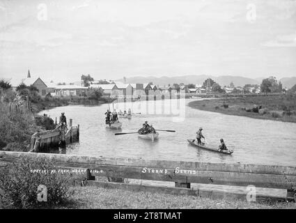 Vista di Opotiki dal molo, vista del fiume Waioeka che guarda a sud dal molo con la cittadina di Opotiki in lontananza. Tre barche a remi e una canoa scavata sono in primo piano. La barca a remi con il nome "Puhi" dipinto a poppa ha sei bambini Maori a bordo, e due dei ragazzi sono in piedi e tengono i remi in posizione verticale. Un ragazzo Maori è sulla riva sinistra del fiume nei primi anni '1900 Foto Stock