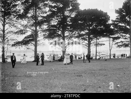 Home Bay, Motutapu Island, foto scattata nel 1910 circa. Vista a est attraverso l'area erbosa pianeggiante dietro la spiaggia verso Waiheke Island in lontananza. La folla cammina nella zona erbosa e si trova sulla spiaggia nelle vicinanze. Un gran numero di yacht e imbarcazioni da diporto sono ancorati nella baia Foto Stock