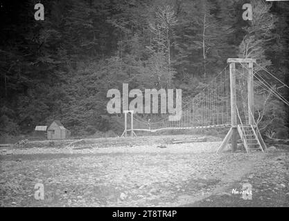 Altalena il ponte sul fiume Maruia con bagni a Maruia Springs visibili sul lato opposto, vista del ponte sospeso sul fiume Maruia, che conduce alle sorgenti di Maruia, con bagni sul lato opposto del fiume. La collina dietro è coperta da un cespuglio nativo risalente al 1910 circa Foto Stock