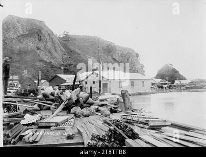 Molo a Whakatane, molo a Whakatane. Ci sono pile di legname in primo piano, diversi uomini stanno sollevando oggetti non identificati, con un mucchio di sacchi pieni dietro di loro. Sullo sfondo centrale si trova l'edificio della New Zealand Loan & Mercantile Agency, con scogliere che si innalzano ripidamente dietro. Fotografato intorno agli anni '1910 Foto Stock