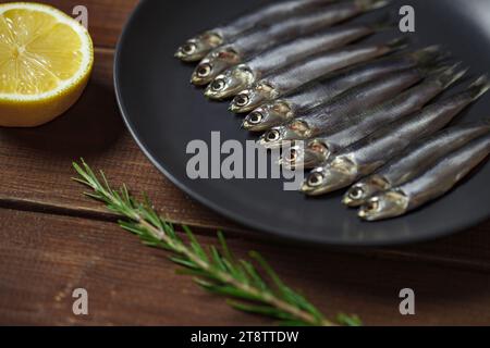 Acciuga europea, hamsa fresca, diversi piccoli pesci disposti in fila su piatto grigio, rosmarino e limone su tavola marrone di legno fondo da tavolo, lato vi Foto Stock