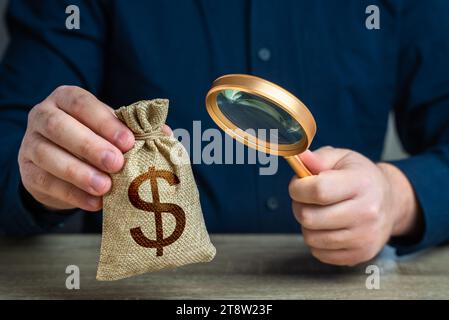 Un uomo esamina un sacco di soldi da un dollaro attraverso una lente di ingrandimento. Indagare sulle origini dei capitali. Antiriciclaggio, evasione fiscale. Termini di studio e condi Foto Stock