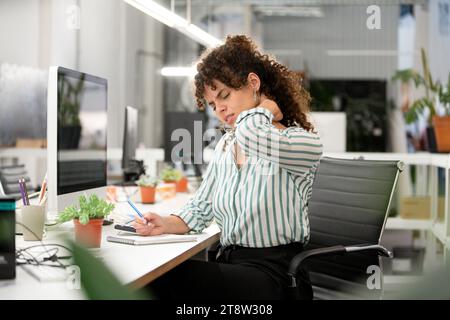 Donna stressata che soffre di contrattura al collo mentre è seduta in ufficio Foto Stock