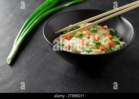 Spaghetti istantanei, cotti con verdure e cipolla verde, in recipiente grigio, bacchette, su sfondo scuro, messa a fuoco selettiva. Foto Stock