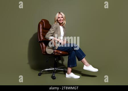 Foto integrale del corpo di una donna del direttore finanziario in banca che aspetta i clienti seduti in poltrona di pelle isolata su sfondo di colore cachi Foto Stock