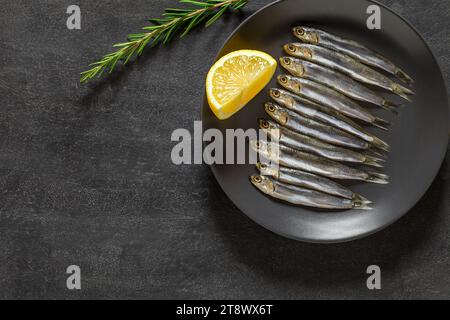 Acciuga europea, hamsa fresca, diversi piccoli pesci su piatto grigio rametto di rosmarino verde, limone, fonte di omega 3, su sfondo scuro, vista dall'alto, spazio t Foto Stock