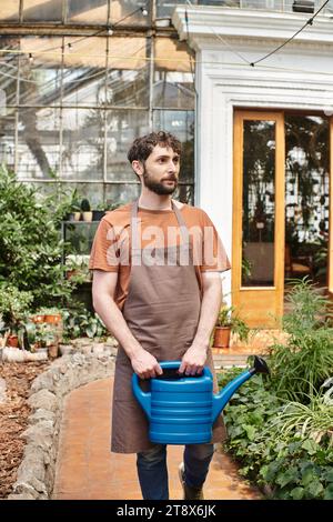 il giardiniere bello e barbuto in grembiule che tiene l'acqua può vicino alle piante verdi in serra Foto Stock