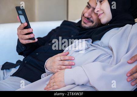 Le coppie che effettuano chiamate online, controllano su Internet la gravidanza nel loro salotto con volti sorridenti Foto Stock