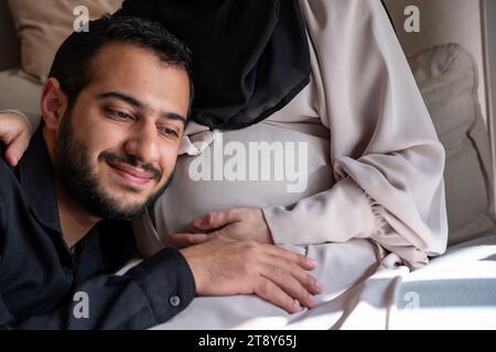 L'uomo arabo sta ascoltando il pancino della sua bella moglie incinta e sorridendo Foto Stock