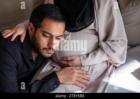 L'uomo arabo sta ascoltando il pancino della sua bella moglie incinta e sorridendo Foto Stock