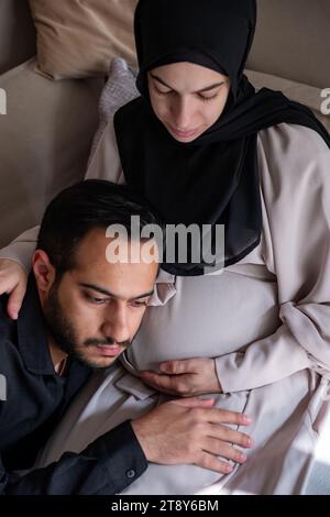 L'uomo arabo sta ascoltando il pancino della sua bella moglie incinta e sorridendo Foto Stock