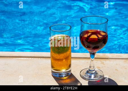 Due bevande rinfrescanti a bordo piscina, un bicchiere di birra e un bicchiere di sangria, sotto il sole luminoso Foto Stock