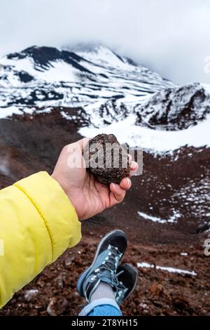 Mano turistica che tiene la pietra lavica sul cratere dell'Etna, Sicilia, Italia. Formazione di roccia lavica vulcanica calda. Paesaggio di crateri pendii ricoperti di neve Foto Stock