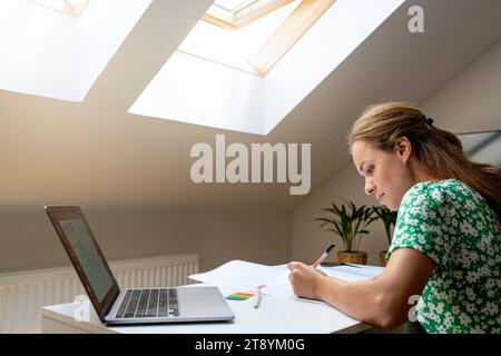 Donna seduto alla scrivania a casa e lavora in remoto utilizzando il suo notebook e documenti cartacei. Foto Stock