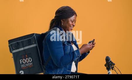 Donna afro-americana che esplora il telefono cellulare mentre è in bicicletta pronta per il servizio di consegna del cibo. Giovane donna di colore che abbraccia l'economia frenetica utilizzando lo smartphone per la navigazione. Foto Stock