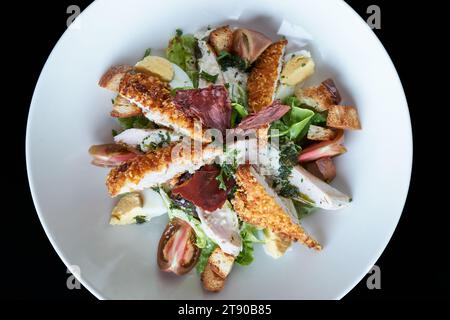 Presentazione dell'insalata Caesar di pollo su un piatto bianco rotondo, vista dall'alto, sfondo nero Foto Stock