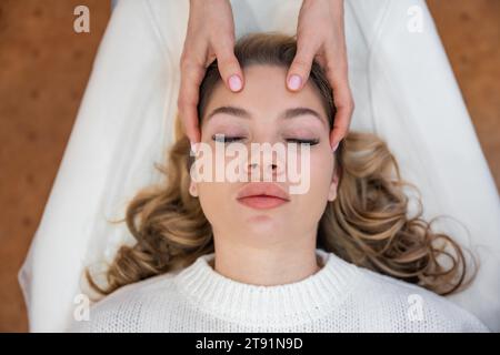 Vista dall'alto delle mani dei guaritori che eseguono la terapia con barre di accesso leggermente toccate sulla testa di una giovane donna, stimolando pensieri ed emozioni di cambiamento positivo Foto Stock