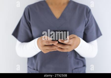 Metà sezione del medico femminile birazziale che indossa scrub usando smartphon in ospedale Foto Stock