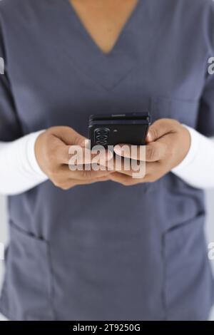 Metà sezione del medico femminile birazziale che indossa scrub usando smartphon in ospedale Foto Stock