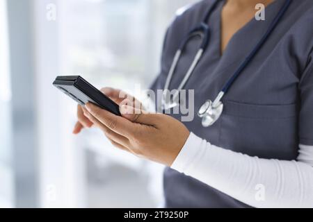 Metà sezione del medico femminile birazziale che indossa scrub usando smartphon in ospedale Foto Stock