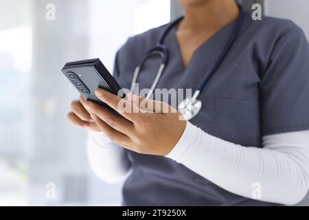 Metà sezione del medico femminile birazziale che indossa scrub usando smartphon in ospedale Foto Stock