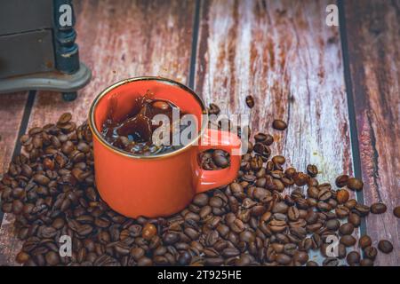 Tazza da caffè rossa con chicchi di caffè che cadono all'interno con effetto spruzzo spruzzare sullo sfondo un macinacaffè manuale Foto Stock