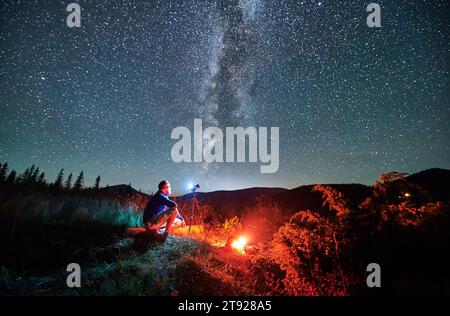 Uomo che scatta foto di cielo stellato con la Via Lattea. Fotografo maschile seduto sul log e fotocamera di messa a fuoco. Uomo con macchina fotografica seduto accanto al fuoco del campo in montagna. Foto Stock