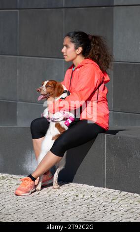 Una giovane donna, vestita di abbigliamento sportivo, tiene il suo cane nel paesaggio urbano Foto Stock
